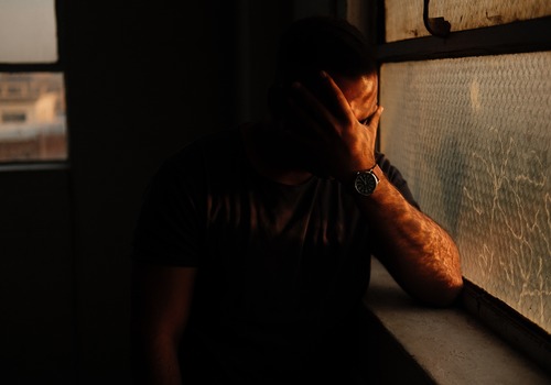man holdding his face in a dark room with sunlight to his left